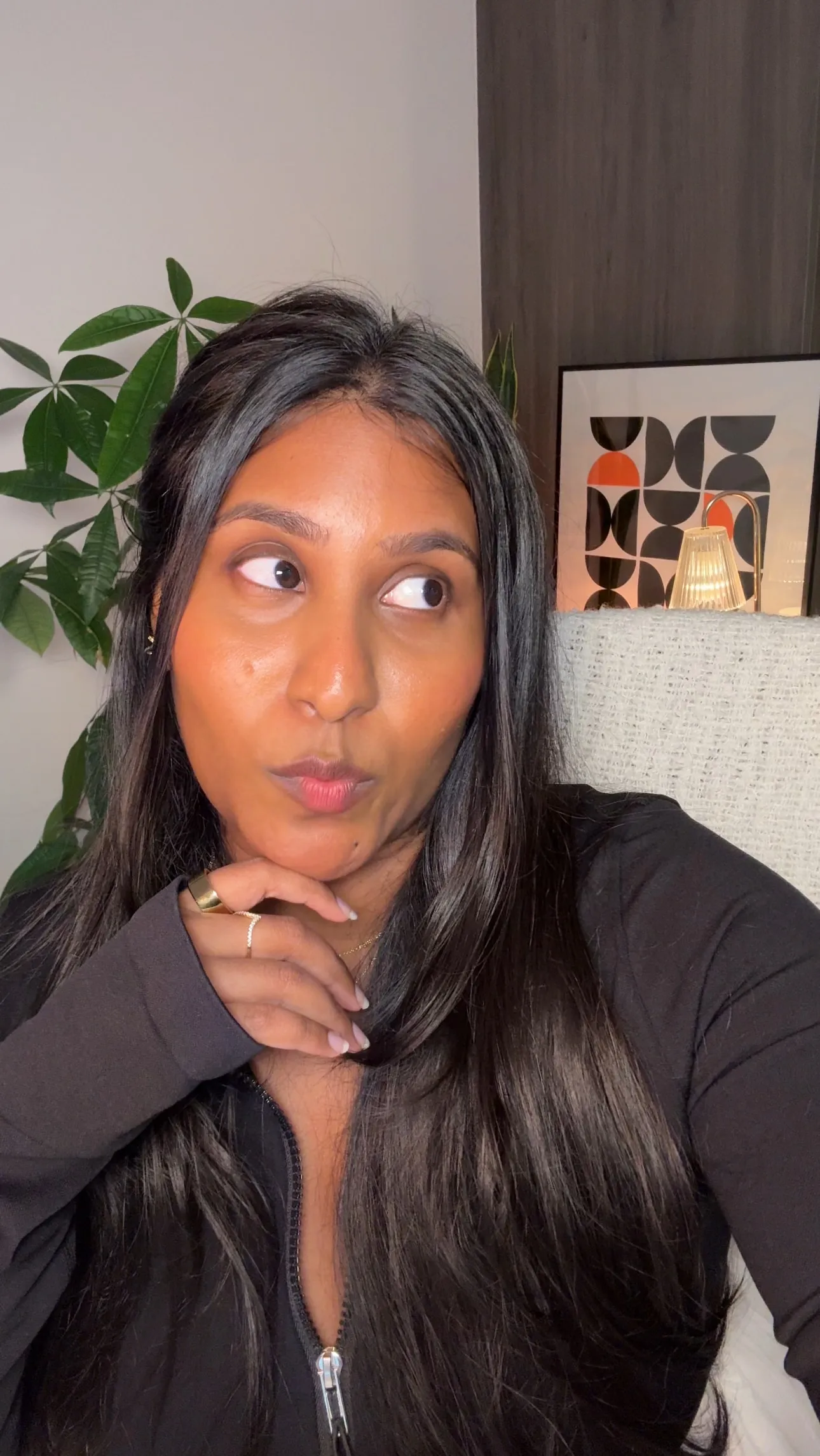 Nina resting her chin on her hand and glancing sideways, a plant and a framed print behind her